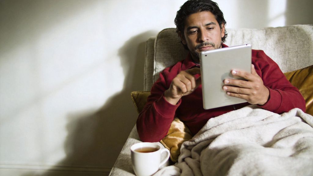 Man relaxing on a sofa with a blanket and tea while reading help articles on a tablet